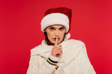 Young woman in santa hat and knitted scarf showing secret gesture isolated on red 