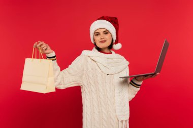 pleased young woman in santa hat and knitted sweater holding laptop and shopping bag on red