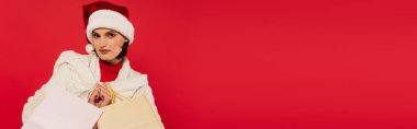 young woman in santa hat and knitted sweater holding shopping bags isolated on red, banner