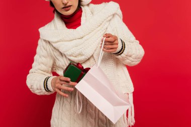 cropped view of young woman in sweater putting gift box with bow into paper bag on red