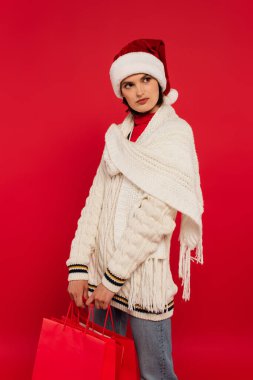 young woman in santa hat and winter sweater holding shopping bags isolated on red