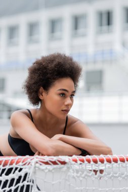 athletic african american woman with crossed arms leaning on sports gates and looking away