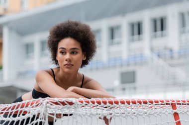 sportive african american woman leaning on sports gates on blurred background