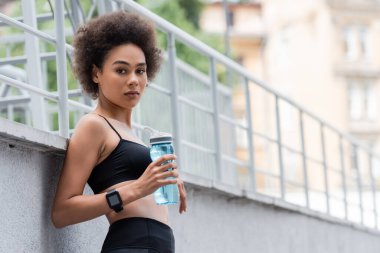 athletic african american woman in fitness tracker holding sports bottle and looking at camera