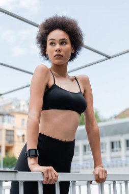 curly african american woman in black sports bra and fitness tracker looking away near fence