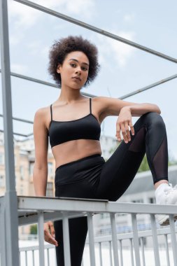 african american woman in black sportswear stepping on fence and looking at camera