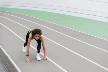 young african american woman in white sneakers standing in low start pose on track
