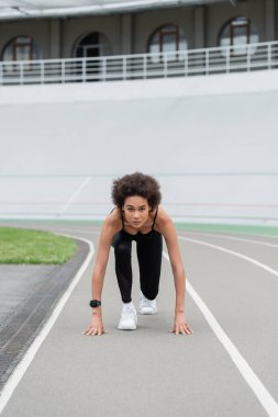 athletic african american woman looking at camera while standing in low start pose on track