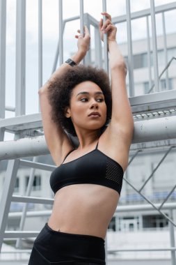 pretty african american woman with slim body posing with raised hands near fence
