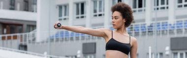 young african american woman in black sports bra training with dumbbell in outstretched hand, banner