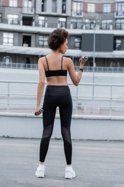 back view of slender african american woman in black leggings exercising with dumbbells