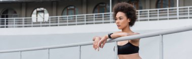 athletic african american woman in black sports bra leaning on fence and looking away, banner