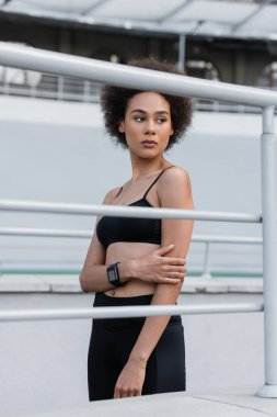 slender african american woman in black sportswear and fitness tracker looking away near fence