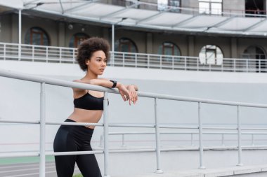 curly african american woman in black leggings and sports bra looking away near fence
