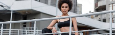 brunette african american woman in black sports bra standing near fence of stadium, banner