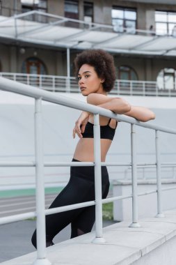 athletic african american woman in black sportswear leaning on fence and looking away