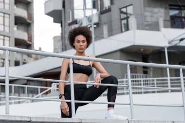 sportive african american woman with curly hair looking at camera on city stadium