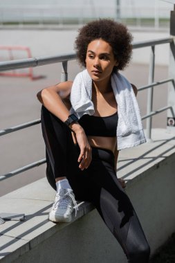 pretty african american sportswoman with towel on neck sitting near fence outdoors