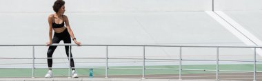 full length of african american woman in black sportswear standing near fence and sports bottle, banner