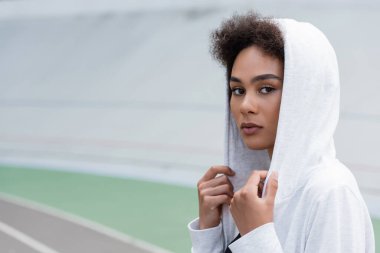 pretty african american sportswoman in white hoodie looking at camera on blurred stadium