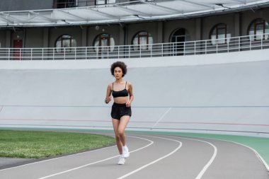 full length of slender african american woman in black sportswear jogging on city stadium