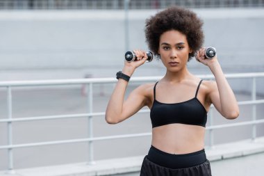 African american sportswoman training with dumbbells and looking at camera on stadium 