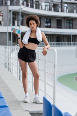 African american sportswoman with towel holding sports bottle near raining of stadium 
