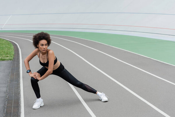 full length of african american woman in black sportswear doing side lunge exercise while stretching on stadium