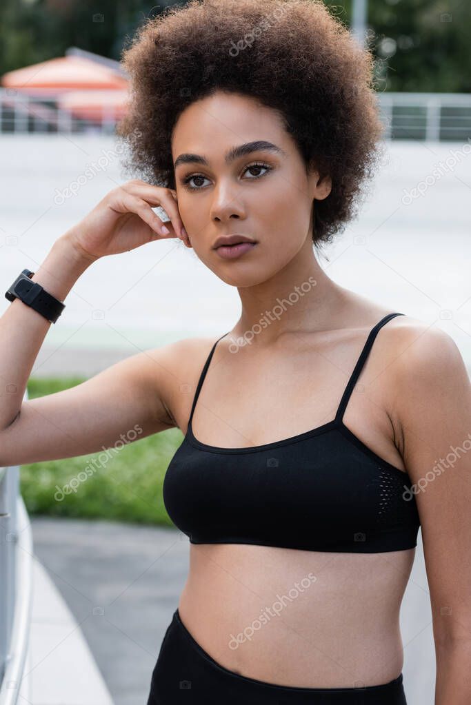 young african american woman in black sports bra and fitness tracker standing with hand near face