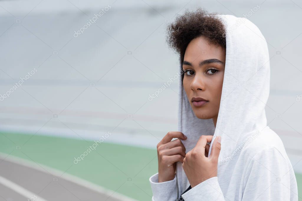 pretty african american sportswoman in white hoodie looking at camera on blurred stadium