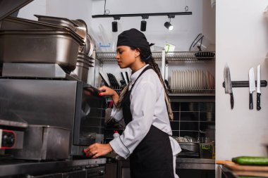 side view of young african american chef in hat using convection oven in kitchen 