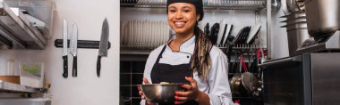 cheerful and young african american chef holding bowl in professional kitchen, banner