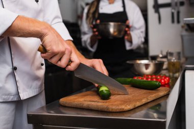 cropped view of chef cutting cucumber on wooden cutting board near blurred colleague 