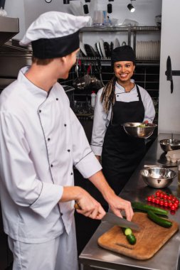happy african american sous chef looking at chef cutting cucumber in kitchen 