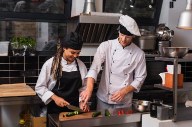 smiling african american sous chef cutting cucumber near head cook in kitchen 