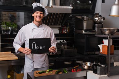happy young chef in uniform holding chalkboard with open lettering in professional kitchen 