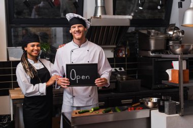happy young chef in uniform holding chalkboard with open lettering near african american colleague in kitchen 