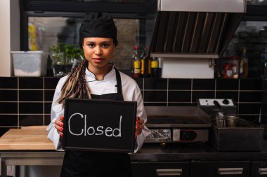young african american chef holding chalkboard with closed lettering in kitchen