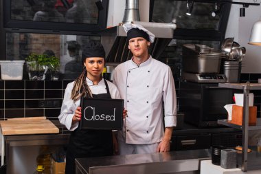 young african american woman holding chalkboard with closed lettering near chef in kitchen