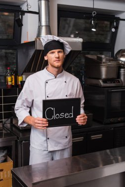 young chef in uniform holding chalkboard with closed lettering in kitchen