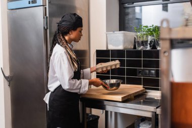 african american chef in apron holding egg carton and bowl in kitchen