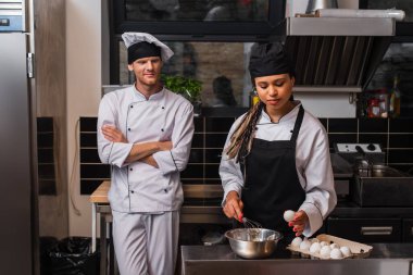 young african american sous chef in apron holding egg near bowl and whisk while cooking near chef in kitchen