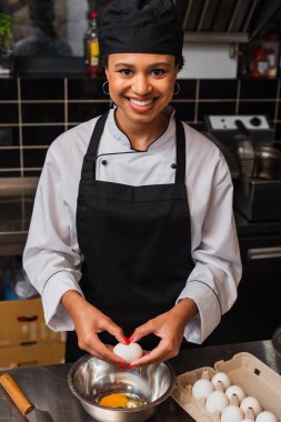 happy african american chef holding raw egg above bowl while cooking in kitchen 