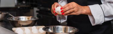 partial view of african american woman braking raw egg above bowl while cooking in kitchen, banner