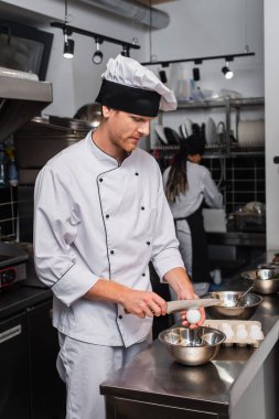 chef in uniform holding knife near raw egg above bowl while cooking in kitchen