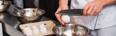 cropped view of chef in uniform holding knife near raw egg above bowl while cooking in kitchen, banner