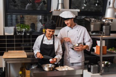 african american sous chef holding knife near raw egg above bowl while cooking near colleague in kitchen