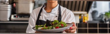 cropped view of african american chef holding plate with freshly cooked salad in professional kitchen, banner