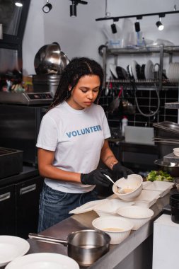 tattooed african american volunteer in t-shirt with lettering holding plastic container with soup and fork in kitchen