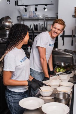 happy interracial volunteers in t-shirts with lettering near plastic containers in kitchen